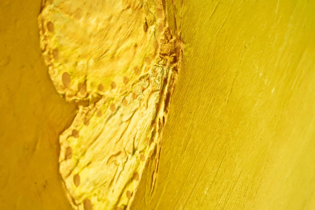Detailaufnahme abstrakter Acrylkunst mit goldener Struktur eines Schmetterlings und leuchtend gelber Oberfläche.